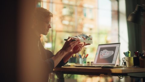 Caucasian Male Artist Redesigning Sneakers, Holding it like a Pieace of Art, Checks 3D Model on Laptop Computer in Creative Loft Environment. Unique Drawing and Conscious Fashion. Slow Motion