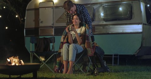 Cinematic shot of young husband is covering with plaid his wife comfortably sitting near their trailer with lighting while enjoying together romantic trip with camping caravan at night.