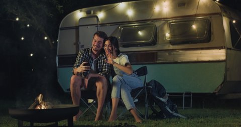 Cinematic shot of carefree couple in love having fun to make video call together to friends with smartphone near their trailer with lighting while enjoying romantic trip with camping caravan at night.