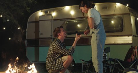 Cinematic shot of young man is making surprise proposal of marriage to his beloved woman near their trailer with lighting while enjoying together romantic trip with camping caravan at night.