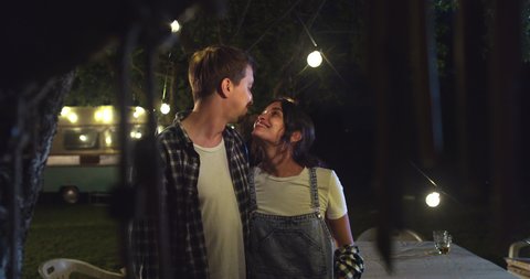 Cinematic shot of happy romantic couple in love enjoying time together and kissing with affection near their trailer with lighting during romantic trip with camping caravan at night.