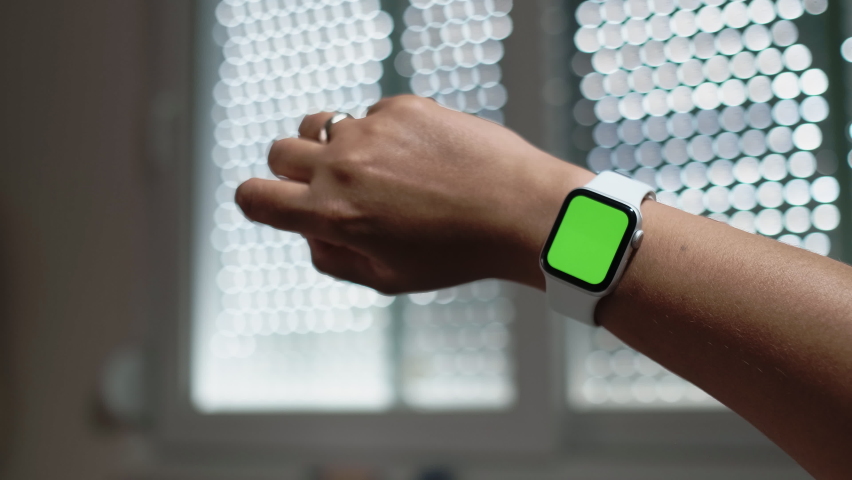 Smart Watch Mock-up or Green screen Luma Key ChromaKey Display on Female Wrist. Wearing Fitness Bracelet with Green Screen Luma Colour Key. Blurred Window Jalousie on Background. Slow Motion Close Up.