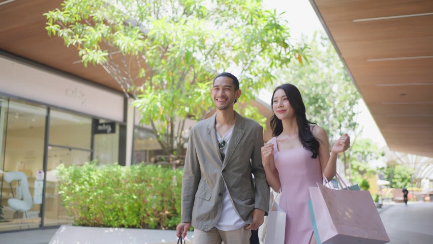 Asian young man and woman shopping goods outdoors in department store. Attractive couple hold shopping bags then walking together with happiness, enjoy purchasing in shopping mall marketplace center. Royalty-Free Stock Footage #1090540491