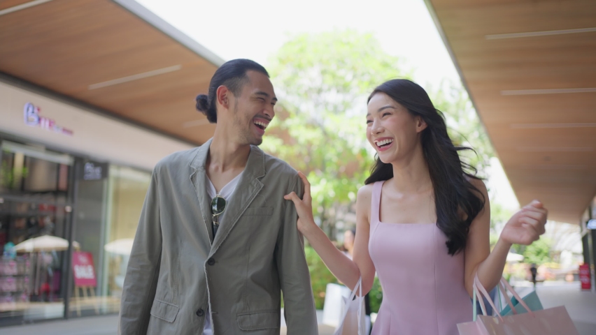 Asian young man and woman shopping goods outdoors in department store. Attractive couple hold shopping bags then walking together with happiness, enjoy purchasing in shopping mall marketplace center. | Shutterstock HD Video #1090540491