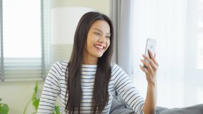 Asian attractive woman using mobile phone video call in living room. Young beautiful girl feeling happy and relax enjoy talking online virtual call on smartphone in house. Technology Lifestyle concept