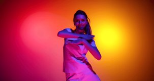 Dance. Tender charming young girl dancing and looking at camera with light smile isolated on yellow red background in neon light, filter. 4K. Concept of emotions, youth, sales and aspiration. Looks