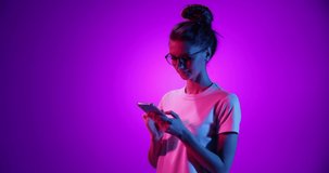 Charming young girl in eyewear using phone, chatting with friend isolated on purple background in neon light, filter. 4K. Concept of emotions, youth, sales and aspiration. Looks happy