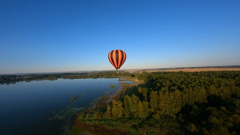 Drone flying close to black and orange hot air balloon floating above lake.