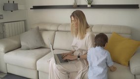 Blonde woman young mother working and raise children. Sitting on sofa in living room and using laptop. Slow motion video footage. Small todler boy back in blue shirt