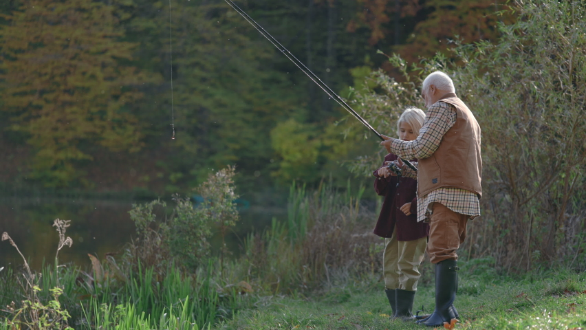 Side view of caucasian senior man standing with his grandson on river bank and showing how to cast spinning rod into lake. Teen boy and his grandfather wearing casual clothes and rubber boots.