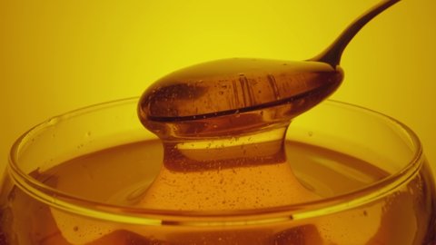 Honey dripping, pouring from spoon in glass. Thick honey molasses dripping into full glass. Close up of golden honey liquid, sweet product of beekeeping. Sugar syrup is pouring on yellow background.