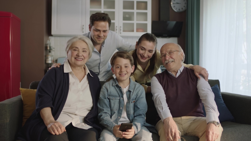 Young couple with children, their son and elderly parents sitting on sofa in living room, taking self portraits together. Portrait of happy cheerful big family smiling at camera in cozy living room. | Shutterstock HD Video #1090548373