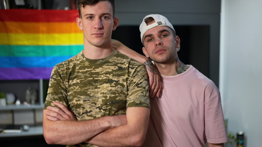 Portrait of excited young gay man boasting engagement ring standing with military boyfriend indoors. Happy Caucasian LGBTQ couple looking at camera bragging proposal with rainbow flag at background