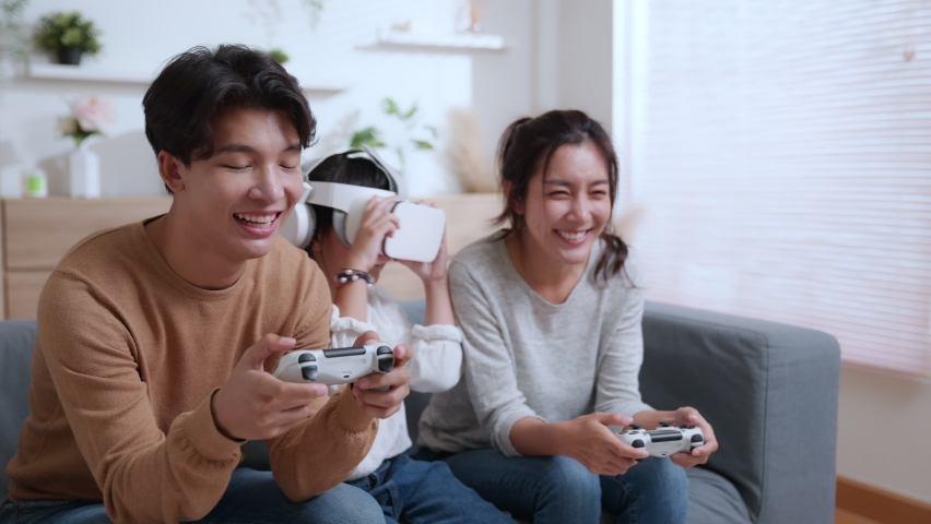 Father and mother playing video game with joystick. A young daughter wearing VR glasses on couch at home. Asian family spending time together on holiday in living room. Attractive happy parents.  | Shutterstock HD Video #1090557189