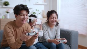 Father and mother playing video game with joystick. A young daughter wearing VR glasses on couch at home. Asian family spending time together on holiday in living room. Attractive happy parents. 