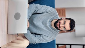 VERTICAL VIDEO POV smiling freelancer Hispanic man sitting on couch in front of laptop. Happy business man portrait working remotely browsing internet relaxing at home posing positive emotion