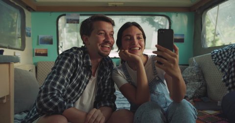 Cinematic shot of carefree couple in love having fun to make video call together to friends with smartphone both sitting inside their trailer while enjoying together romantic trip with camping caravan