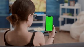 Closeup of young woman holding vertical smartphone with green screen watching social media content in home living room. Girl using touchscreen mobile phone with chroma key looking at screen.