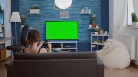 Gamer girl playing console video games with wireless controller on green screen tv relaxing on couch in living room. Young woman enjoying free time at home doing online gaming on chroma key display.