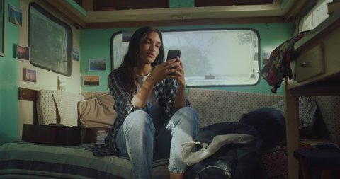 Cinematic shot of young woman is having fun to use smartphone for navigation in internet and communicate with friends on social media inside trailer during family trip with rv camping caravan.