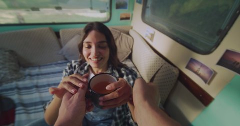 Cinematic authentic pov of young husband is bringing fresh orange juice to his wife and caressing her with affection inside their trailer while enjoying together romantic trip with camping caravan.