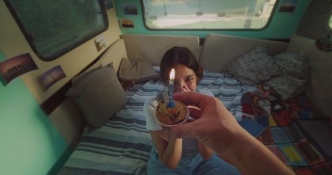 Cinematic authentic pov of young husband is bringing muffin with candle to wife as surprise to celebrate her birthday inside their trailer while enjoying together romantic trip with camping caravan.