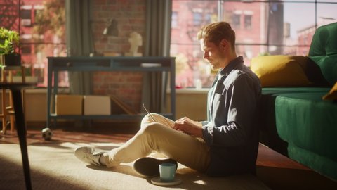 Handsome Young Man Using Laptop, Working from Home Living Room with Big Windows. Male Sitting on Floor, Using Computer, Writing Project Plan. Successful Manager Doing Remote Work.
