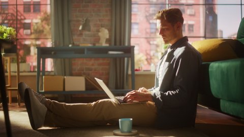 Young Man is Sitting on a Floor, Working or Studying on Laptop Computer. Cozy Loft Living Room with Big Windows, Modern Interior, Green Sofa and Wooden Flooring.