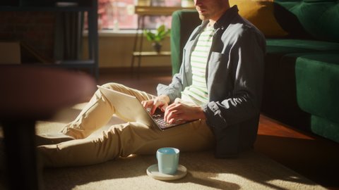 Portrait of an Attractive Young Adult Male Sitting on a Floor and Using Laptop Computer in Sunny Loft Living Room. Successful Man Working from Home in Bright Apartment.