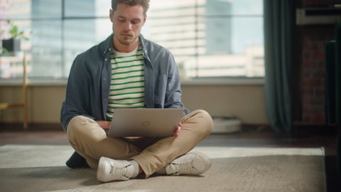 Handsome Young Man Using Laptop, Working from Home Living Room with Big Windows. Male Sitting on Floor, Using Computer, Writing Project Plan. Successful Manager Doing Remote Work.