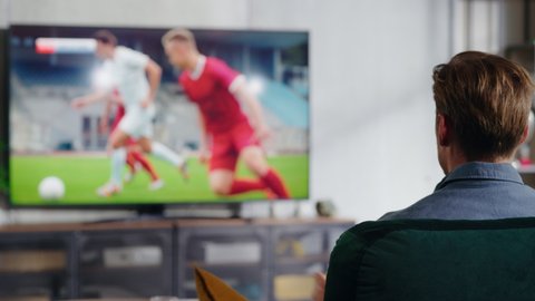 Male Soccer Fan Relaxes on a Couch, Watch a Sports Match at Home in Stylish Loft Apartment. Excited Young Man Cheer for Their Favorite Football Club, Celebrate Player Scoring a Goal.