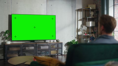 Young Handsome Man Sitting on a Sofa and Watching TV with Horizontal Green Screen Mock Up. It's Day Time in Room at Home Living Room in Loft Apartment.