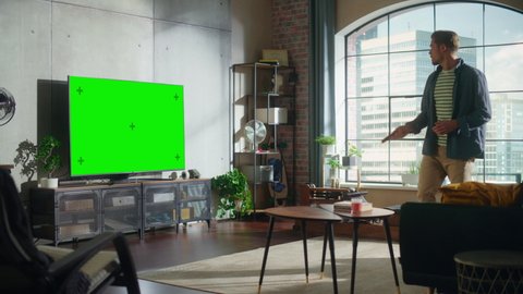 Young Handsome Man Sitting Down on a Sofa and Watching TV with Horizontal Green Screen Mock Up. It's Day Time in Room at Home Living Room in Loft Apartment.
