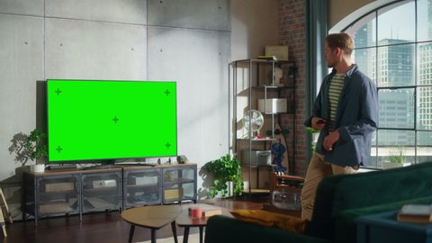 In Loft Living Room an Attractive Man Sitting Down on a Couch, Turns On TV with Green Screen Mock Up Display with a Controller. Male Enjoys Leisure Time at Home.