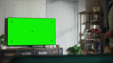 Young Handsome Man Spending Time at Home, Sitting Down on a Couch and Turning On TV with Green Screen Mock Up Display in Stylish Loft Apartment. Man Stream Movie at Home.