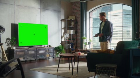 Young Handsome Man Spending Time at Home, Sitting Down on a Couch and Turning On TV with Green Screen Mock Up Display in Stylish Loft Apartment. Man Stream Movie at Home.