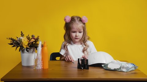 A little girl on a yellow background is sitting at a table with a manicure set. A girl in a beauty salon does a manicure. Children are like adults.