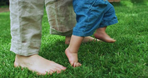 Authentic close up shot of little toddler baby boy feet taking his first steps with mother's help on green lawn in sunny day.