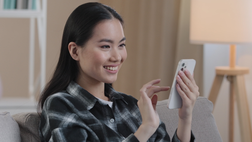 Smiling happy Asian woman typing swipe touchscreen of phone Korean girl cheerful choosing shopping in online e-commerce app female enjoy mobile website play game at home follow post in social media Royalty-Free Stock Footage #1090574169