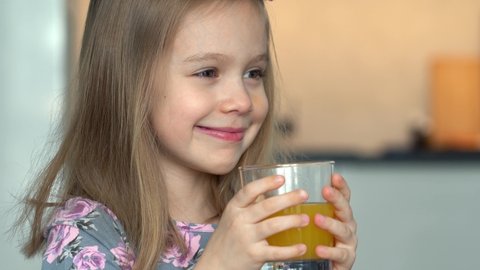 Close up of little girl laughs and fun drinks orange juice. Happy child drinks juice on kitchen at home