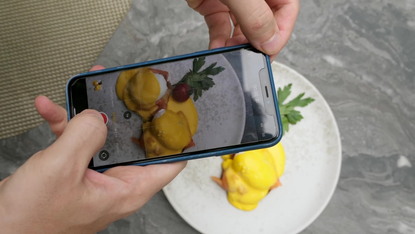 Man making vertical video, stories, photo for social media of delicious restaurant food on smartphone. Male influencer shoot clip, film, reels on phone camera of eggs benedict breakfast. Close-up.