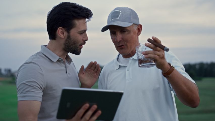 Smiling men calling video holding tablet on golf course. Two golfers introduce partner at country club. Close up rich old senior drink glass whiskey smoking cigar on online meeting. Wealthy concept.