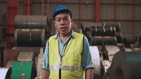 Happy Asian man engineer in uniform wearing blue safety helmet and glove waving hand and talking by video call at warehouse in factory. Senior male worker smile looking camera and chatting on video. 