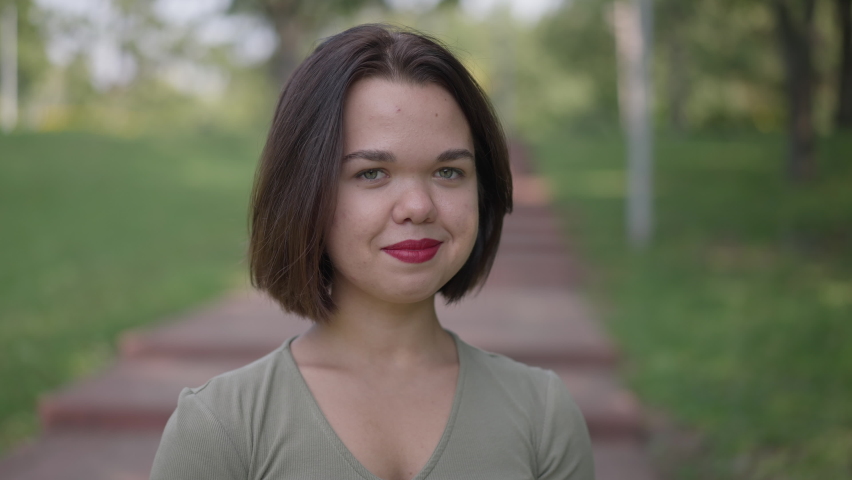 Close-up coquette Caucasian little woman gesturing air kiss smiling looking at camera. Portrait of confident happy positive young person with dwarfism posing in slow motion flirting standing in park