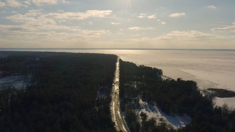 4k drone aerial footage icy road with car traffic between fluffy winter forest and frozen lake. Kiev Sea, people came to have fun on the ice on winter weekends. Sunny weather, blue sky, shiny ice road