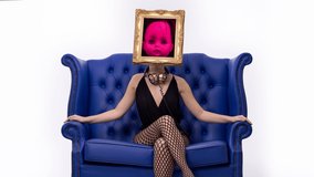 Amazing female with a gold picture frame as a head sitting in a transparent chair. On the screen is a childs doll head moving
