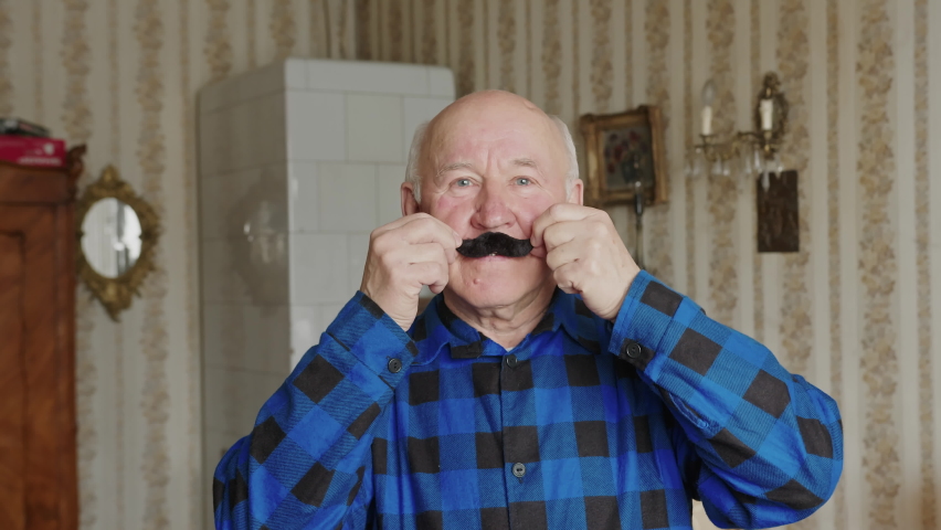 playful senior Caucasian man holding a fake mustache in the living room medium closeup senior people concept. High quality 4k footage