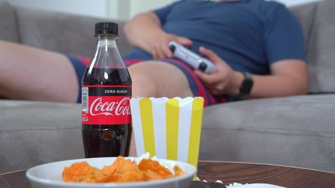 Budva, Montenegro - April 21, 2022: Man is sitting on the couch with gamepad in his hand playing video games eating chips and popcorn and drinking Coca Cola. Dependence to video games and fast food
