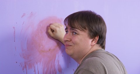 Mischievous plump man caught smearing pink paint on purple wall after renovation. Young guy turns and shrugs feeling guilty close view