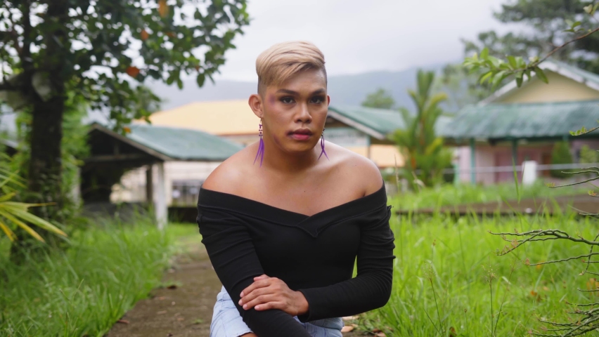 A Lonely Gay With Few Bruises On Face Wearing Black Off-Shoulder Shirt And Feather Earrings, Sitting On Pathway Looking To The Camera In A Rural Farm In Philippines. - Medium Closeup Shot Royalty-Free Stock Footage #1090589211
